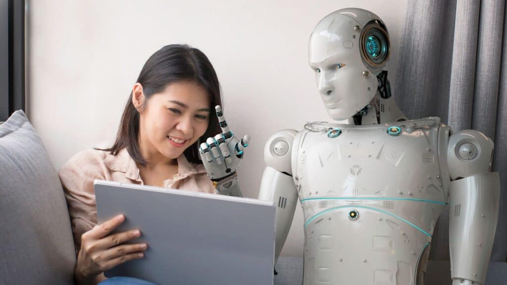 A robot sits next to a woman on a couch while they look at a computer together 