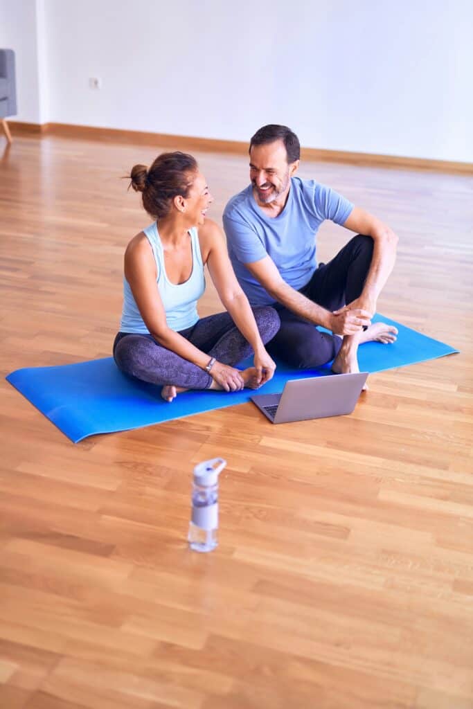 A health coaching couple sit together on a yoga mat smiling at each other with their computer in front of them