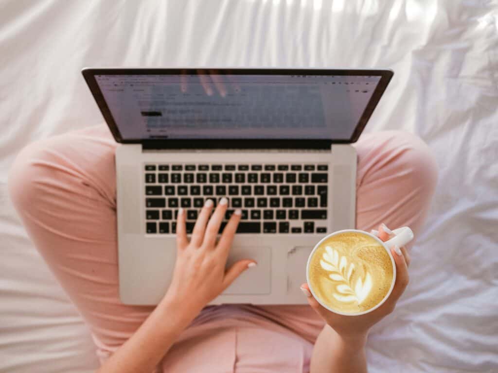 a health coach sits on her bed with her legs crossed, laptop in lap, and coffee in hand 